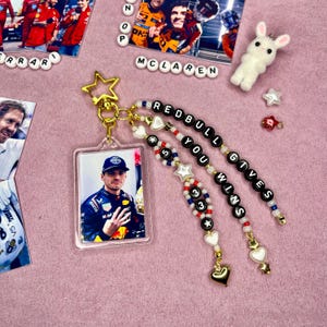 May include: A clear acrylic keychain featuring a photo of a race car driver, with a gold star charm. The keychain is adorned with beaded accents spelling "REDBULL YOU WINS". Additional charms include stars and hearts.