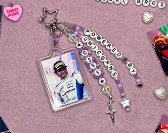 Puede incluir: Un llavero acrílico transparente con una foto de un hombre con uniforme de carreras. El llavero tiene una parte superior plateada en forma de estrella y detalles de cuentas en morado y blanco. Las palabras "POWERED BY UNICORNS S5" están escritas en las cuentas. Un dije rosa en forma de corazón dice "SWEET HEART".