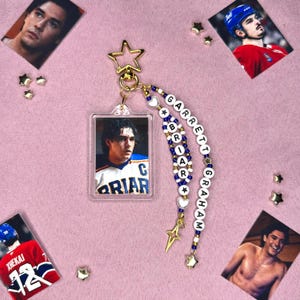 May include: A clear acrylic keychain featuring a photo of a hockey player in a jersey. The keychain is adorned with a gold star-shaped charm and a beaded strand spelling out "GARRETT BRIAR GRAHAM". Additional photos and star-shaped embellishments are scattered around.