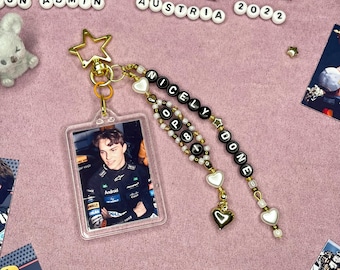 May include: A clear acrylic keychain featuring a photo of a person in a racing suit. The keychain has gold-colored hardware, a star charm, and beaded accents spelling "NICELY DONE". Additional beads are white and black.