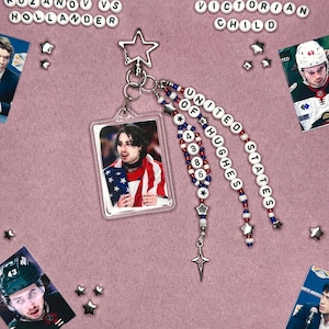 Puede incluir: Un llavero con un colgante rectangular transparente con una foto de una persona envuelta en una bandera estadounidense. El llavero tiene hebras de cuentas con las palabras "UNITED OF HUGHES STATES" y dijes de estrella.