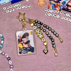 May include: A keychain with a clear plastic photo card featuring a race car driver. The keychain has three beaded strands with black and white letter beads spelling "NOT BAD FOR A NO 2 DRIVER" and gold beads with white stars.