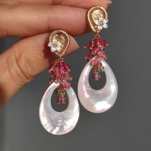 May include: Elegant gold-toned earrings featuring pearlescent, teardrop-shaped pendants. Each earring has a cluster of pink and green gemstones, and a small white flower. The earrings are designed with a delicate, wire-wrapped detail.