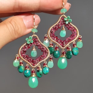 Op de afbeelding: Goudkleurige oorhangers met een filigraan ontwerp. De oorbellen hebben een centraal patroon van kleine rode kralen, geaccentueerd met groene en turquoise druppelvormige stenen. Kleine groene bloemaccenten zijn ook aanwezig.