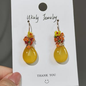 May include: A pair of gold-toned drop earrings with a teardrop-shaped yellow stone. The earrings are adorned with small orange, red, and yellow beads. The card behind the earrings reads "Ukuly Jewelry" and "Thank You".