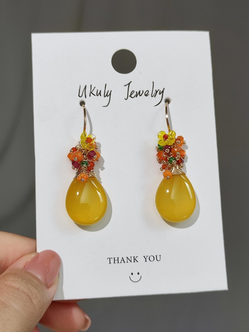 May include: A pair of gold-toned drop earrings with a teardrop-shaped yellow stone. The earrings are adorned with small orange, red, and yellow beads. The card behind the earrings reads "Ukuly Jewelry" and "Thank You".