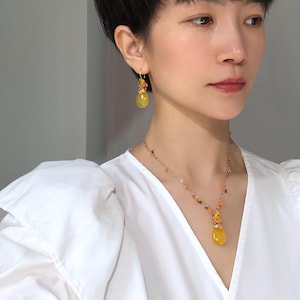 May include: A woman wearing a yellow gemstone jewelry set, including a necklace and earrings. The necklace features a teardrop-shaped pendant and small, colorful beads. The earrings have a similar design, with a cluster of colorful beads and a larger yellow stone.