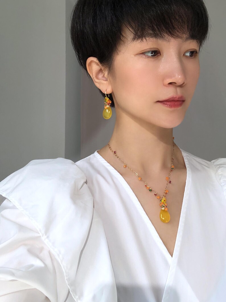 May include: A woman wearing a yellow gemstone jewelry set, including a necklace and earrings. The necklace features a teardrop-shaped pendant and small, colorful beads. The earrings have a similar design, with a cluster of colorful beads and a larger yellow stone.