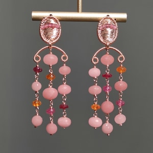 Peut inclure: Boucles d'oreilles artisanales composées de fils cuivrés et de pierres précieuses roses et oranges. Elles présentent des ovales en haut et plusieurs rangées de perles roses et oranges, créant un effet pendant. Les boucles sont suspendues à une barre dorée.