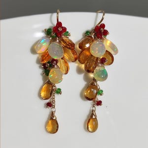 Peut inclure: Une paire de boucles d'oreilles pendantes dorées. Chaque boucle d'oreille présente des grappes de pierres précieuses opalescentes, ambrées et jaune, accentuées de perles rouges en forme de fleurs et de perles vertes. Les boucles d'oreilles ont un aspect délicat et artisanal.