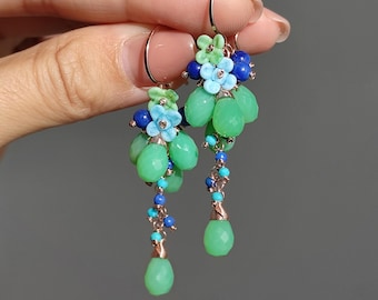 Puede incluir: Pendientes colgantes con cuentas verdes, azules y turquesas. Los pendientes tienen un diseño floral con pequeñas flores azules y verdes. Cuelgan de aros de color dorado.