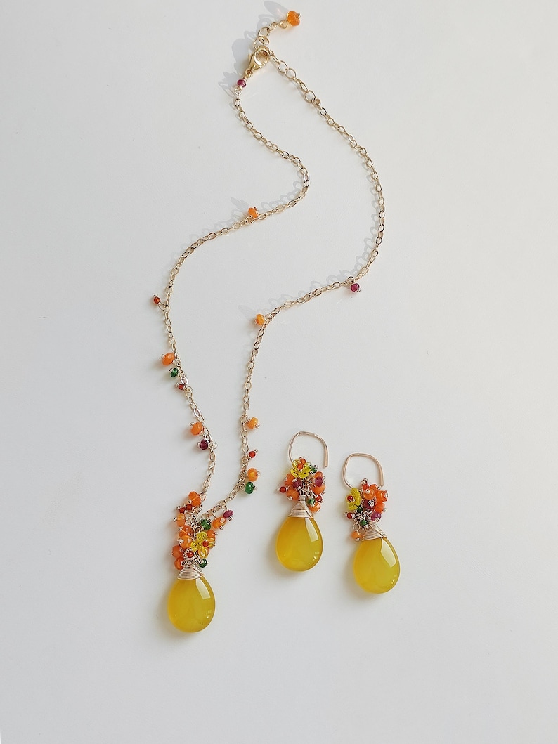 May include: A gold-toned necklace and matching earrings set. The necklace and earrings feature yellow teardrop-shaped pendants and clusters of colorful beads. The jewelry is set against a white background.
