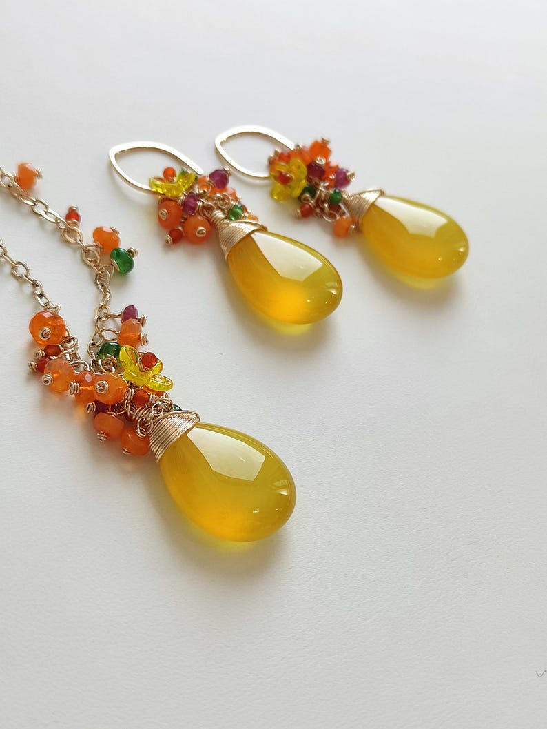 May include: A jewelry set featuring a necklace and earrings. The necklace and earrings have a teardrop-shaped, yellow gemstone pendant. The pendants are accented with colorful beads and wire wrapping. The earrings have gold-colored ear wires.