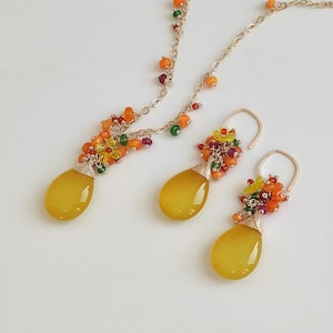 May include: A gold-toned jewelry set featuring a necklace and earrings. Each piece has a large, yellow, teardrop-shaped stone. The necklace and earrings are adorned with clusters of small, colorful beads in shades of orange, green, and red.