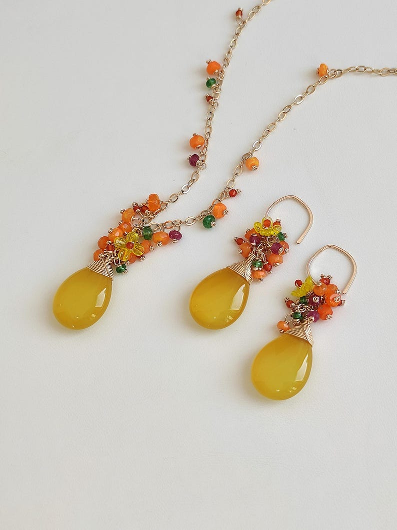 May include: A gold-toned jewelry set featuring a necklace and earrings. Each piece has a large, yellow, teardrop-shaped stone. The necklace and earrings are adorned with clusters of small, colorful beads in shades of orange, green, and red.