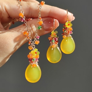 May include: A gold-toned jewelry set featuring a necklace and earrings. The necklace and earrings have a teardrop-shaped yellow stone pendant. The necklace and earrings are adorned with colorful beads and small flower accents.
