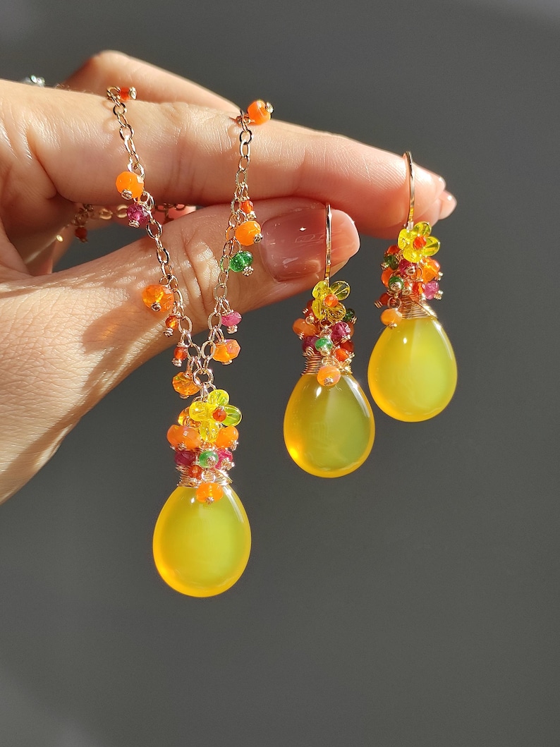 May include: A gold-toned jewelry set featuring a necklace and earrings. The necklace and earrings have a teardrop-shaped yellow stone pendant. The necklace and earrings are adorned with colorful beads and small flower accents.