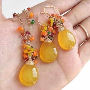May include: A gold-toned necklace and matching earrings set. The jewelry features teardrop-shaped yellow gemstones, accented with clusters of orange, yellow, and green beads. The earrings have a hook closure. The necklace has a delicate chain.