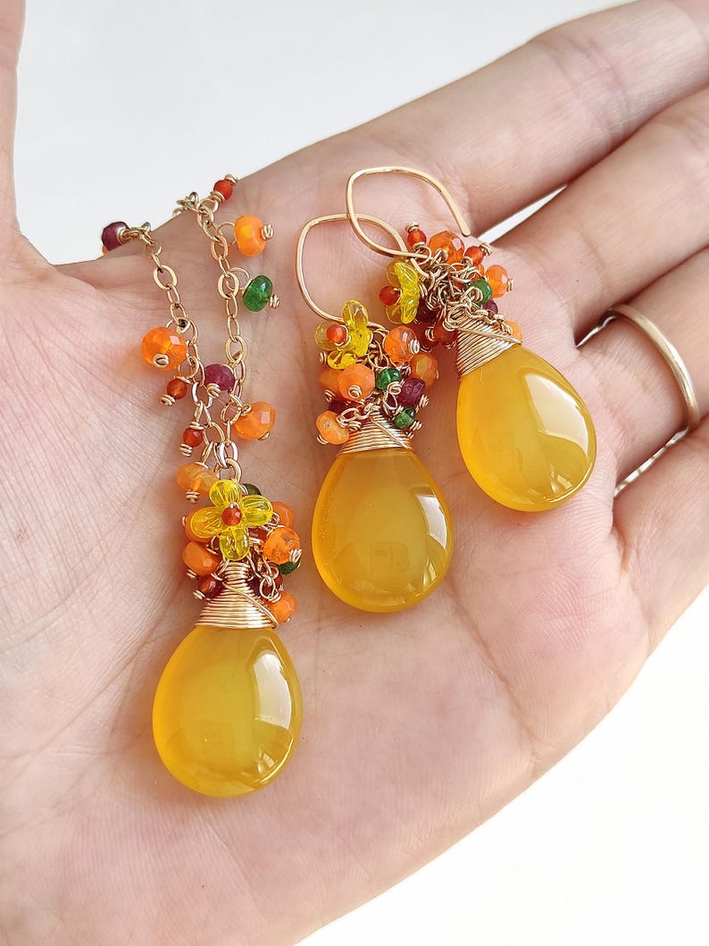May include: A gold-toned necklace and matching earrings set. The jewelry features teardrop-shaped yellow gemstones, accented with clusters of orange, yellow, and green beads. The earrings have a hook closure. The necklace has a delicate chain.