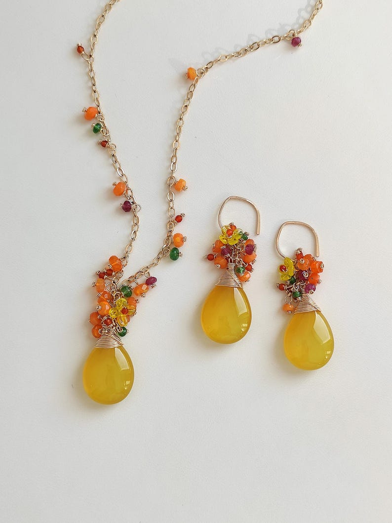 May include: A gold-toned necklace and matching earrings set. The necklace and earrings feature teardrop-shaped yellow gemstones, accented with colorful beads. The gemstones are wire-wrapped, and the jewelry is set against a white background.