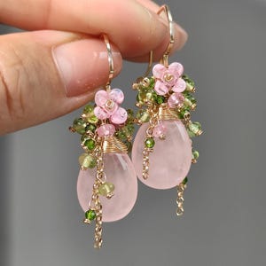 Può includere: Un paio di orecchini color oro con pietre rosa a forma di goccia, impreziositi da piccoli grappoli di fiori rosa e perline verdi. Delicate catene dorate pendono dalle pietre, creando un design elegante e femminile.