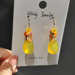 May include: A pair of dangle earrings with yellow teardrop-shaped stones. The earrings have a cluster of small, colorful beads and yellow flower accents at the top. The card behind the earrings reads "Ukuly Jewelry" and "THANK YOU".