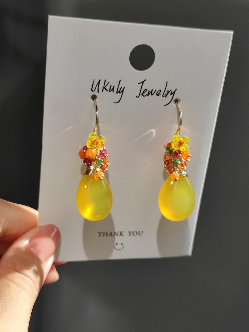 May include: A pair of dangle earrings with yellow teardrop-shaped stones. The earrings have a cluster of small, colorful beads and yellow flower accents at the top. The card behind the earrings reads "Ukuly Jewelry" and "THANK YOU".