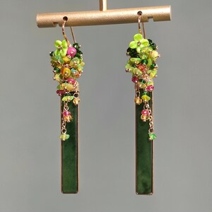 Può includere: Un paio di orecchini pendenti con pendenti rettangolari verde scuro. Gli orecchini presentano grappoli di perline colorate in verde, giallo, rosa e rosso. Sono sospesi a ganci color oro, mostrando un design vibrante e artistico.