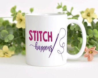 May include: White ceramic mug with the text "STITCH happens" in pink and purple glitter lettering. A purple needle and thread design is on the right side of the mug. The mug has a white handle and is surrounded by greenery.
