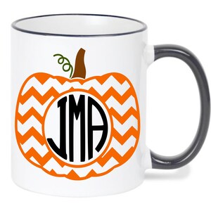 May include: White ceramic mug with a black handle and rim. The mug features a large orange pumpkin design with a chevron pattern. Inside the pumpkin is a white circle with the initials "JMA" in black.