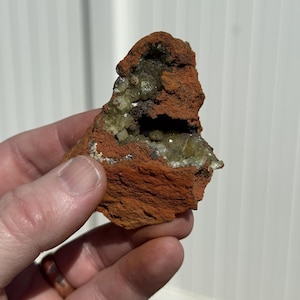 May include: A rough, reddish-brown rock formation with a cavity revealing a cluster of small, light green crystals. The rock is held in a hand, showcasing its natural texture and color variations. The overall appearance suggests a mineral specimen.