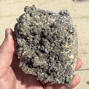 Può includere: Una mano che tiene una formazione rocciosa ruvida e grigia con una superficie strutturata. La roccia presenta piccoli cristalli scintillanti e un mix di tonalità grigio scuro e chiaro. L'aspetto generale suggerisce un campione minerale naturale.