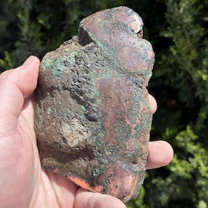 May include: A rough, irregularly shaped rock with a mix of copper and green hues. The surface has a textured appearance, with areas of shiny copper and patches of green oxidation. The rock is held in a hand, showcasing its natural, raw form.
