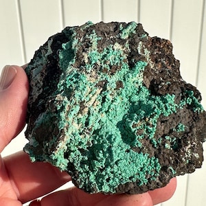 Op de afbeelding: Een ruwe, donkergrijze rots met een groene minerale coating. Het mineraal lijkt in clusters te zitten en heeft een licht iriserende glans.