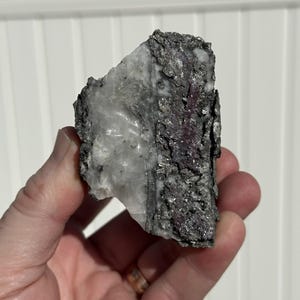 May include: A rough-hewn mineral specimen with a mix of colors and textures. The rock features a translucent white section, contrasted by areas of dark gray and patches of a reddish-purple hue. The surface is uneven and appears to be naturally formed.