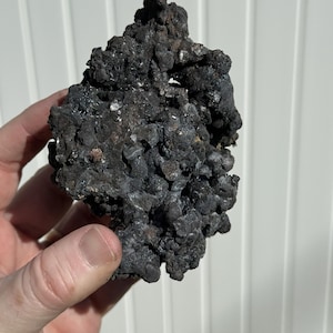 May include: A dark, textured mineral specimen with a rough, uneven surface. The rock is primarily dark gray and black, with some areas appearing metallic. The mineral is held in a hand against a white background.