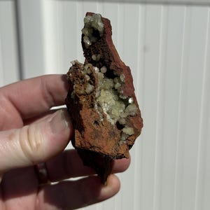 May include: A reddish-brown mineral specimen with clusters of small, pale green crystals. The mineral is held in a hand, showcasing its natural texture and colour variations. The specimen has a rugged, organic shape.