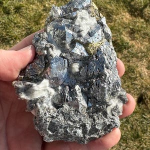 Può includere: Un gruppo di cristalli color argento incastonati in una roccia grigia. I cristalli hanno un bagliore metallico e sono disposti in modo casuale. La roccia è ruvida e testurizzata.