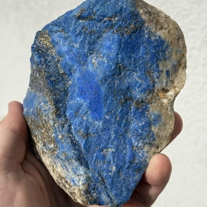 May include: A rough, natural lapis lazuli stone with a deep blue color and flecks of gold. The stone has a textured surface and is held in a hand.