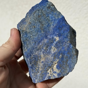May include: A hand holding a rough-cut lapis lazuli stone. The stone is a deep blue color with flecks of gold and white throughout. The surface is textured and uneven, showcasing the natural beauty of the mineral. This raw gemstone is a unique addition to any collection.