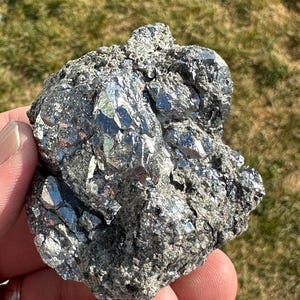Può includere: Un grande masso metallico di colore argento, ruvido e con una struttura cristallina. La roccia ha una superficie ruvida e irregolare ed è ricoperta da piccoli cristalli lucenti.