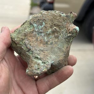 May include: A rough, irregularly shaped rock with a mix of brown and green hues. The surface has a textured appearance, with patches of green suggesting the presence of copper minerals. The rock is held in a hand, showcasing its size and form.