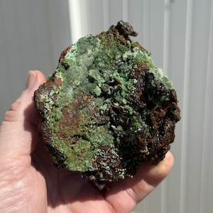 May include: A rough, natural mineral specimen with a mix of colors. The rock features areas of deep brown, reddish-brown, and vibrant green, with a textured, uneven surface. The mineral appears to be held in a hand.