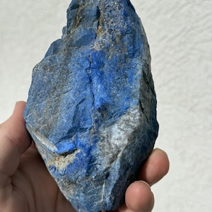 May include: A rough, natural lapis lazuli stone with deep blue and gray hues. The stone has a textured surface with visible crystalline structures. The lapis lazuli is held in a hand, showcasing its size and natural beauty.