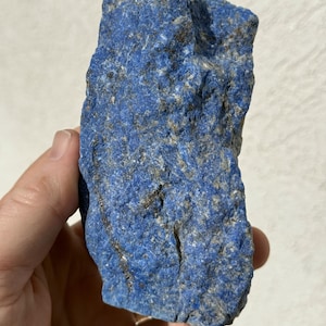 May include: A rough, natural lapis lazuli stone. The deep blue stone has flecks of gold and gray throughout its textured surface. The stone is held in a hand against a white background.