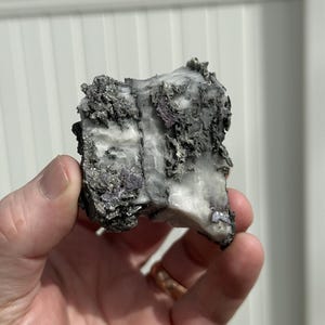 Może przedstawiać: Dłoń trzymająca surowy, naturalny okaz minerału. Kamień charakteryzuje się mieszanką szarych, białych i fioletowych odcieni, o teksturowanej, nierównej powierzchni. Minerał wydaje się być surowym, nieoszlifowanym kamieniem.