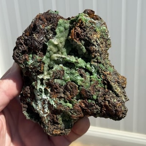 May include: A rough, natural mineral specimen featuring a mix of textures and colors. The rock has a dark brown base with areas of vibrant, light green crystalline formations. The overall appearance is rugged and earthy, showcasing the raw beauty of natural minerals.
