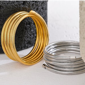 May include: Two coiled bracelets. One is gold-toned, the other is silver-toned. Both are made of a flexible, snake-like material. The gold bracelet is coiled around itself, while the silver bracelet is partially uncoiled, revealing a magnetic clasp.