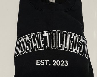 Puede incluir: Sudadera negra con texto bordado en blanco que dice "COSMETOLOGIST EST. 2023".