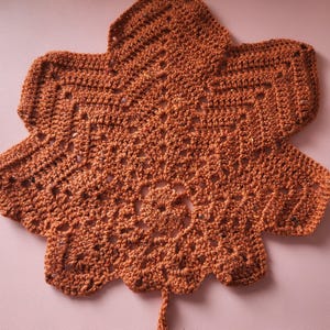 Peut inclure: Napperon en forme de feuille, fait à la main, en crochet marron. Le napperon présente un motif ajouré détaillé avec un motif floral central et des bords festonnés. La forme de la feuille est clairement définie, avec une tige qui s'étend du bas.